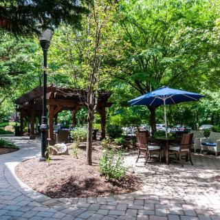 A peaceful garden setting with outdoor seating under a blue umbrella, surrounded by trees and a brick pathway.