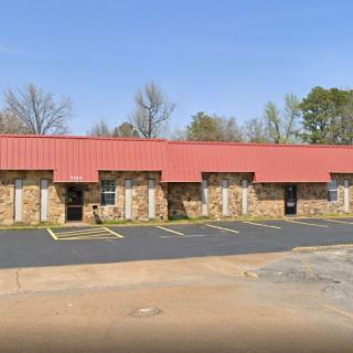 A row of commercial buildings with a red roof and brick facade, featuring an empty parking lot on a clear day.