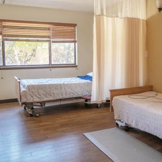 A hospital room with two beds separated by a curtain, featuring large windows and wooden flooring.