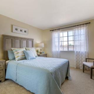 A cozy bedroom with a bed covered in light blue bedding, softly lit by a window draped with sheer curtains.
