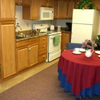 A cozy kitchen and dining area featuring wooden cabinetry, a white refrigerator, oven, and a microwave. A small round table with a red and blue tablecloth is set for dining.