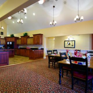 A spacious dining area with wooden furniture and a connected kitchen, featuring warm lighting and a carpeted floor.