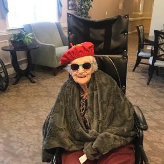 An elderly person sitting in a wheelchair indoors, wearing a red hat and sunglasses with a cheerful expression.