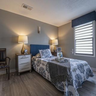 A modern bedroom featuring a single bed with floral bedding, flanked by two nightstands and lamps, under a window with blinds.