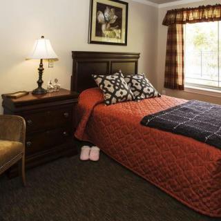 A cozy bedroom with a warmly decorated bed, nightstand, and lamp by a window.