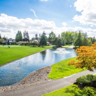 A scenic view of a park with a lake, surrounded by trees and a pathway under a partly cloudy sky.