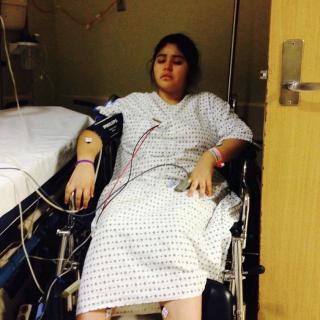 A patient in a hospital gown seated in a wheelchair connected to medical equipment.