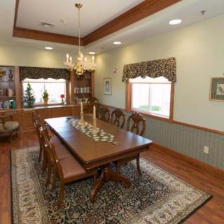 A traditional dining room with a large wooden table, surrounded by chairs and lit by a chandelier. The room features wood flooring, elegant curtains, and decorative elements.