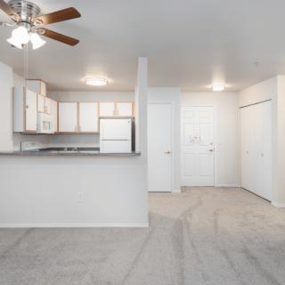 Bright and open apartment space with a kitchen and living area, featuring white walls and carpet flooring.
