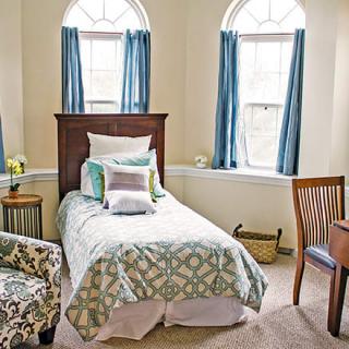 A cozy bedroom with a single bed, adorned with decorative pillows, next to large windows with blue curtains. An armchair and wooden desk complete the setup.