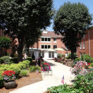 A serene garden courtyard with lush greenery, vibrant flowers, and outdoor seating leading to a multi-story brick building.