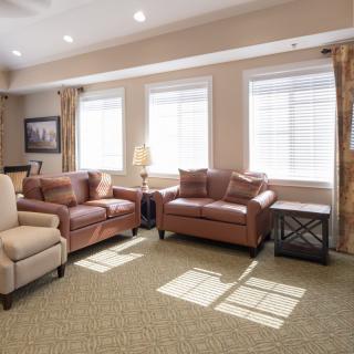 A cozy living room with sofas and a carpet, lit by natural light from large windows.