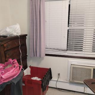 A cozy room featuring a dresser, window with curtains, and an air conditioner, alongside some clothing and storage baskets.