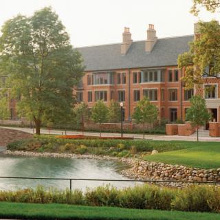 A serene campus scene featuring a large brick building next to a pond surrounded by lush greenery.
