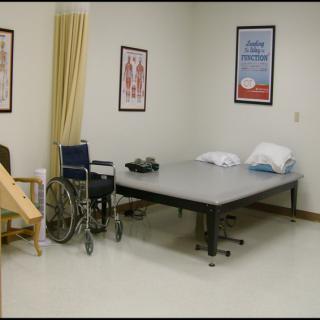 A medical examination room with a wheelchair and examination table, featuring anatomical posters on the walls.