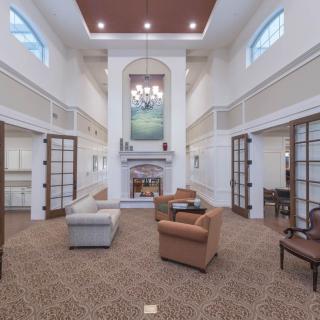 A spacious living room with a high ceiling, featuring a seating area around a central fireplace and elegant chandeliers.