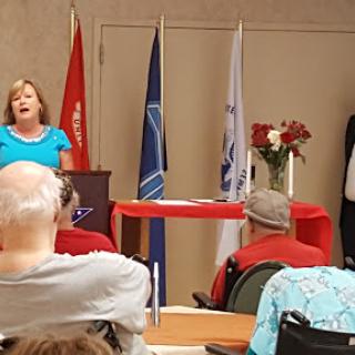 A speaker delivers a presentation at a community event, with an audience gathered and various flags displayed.