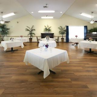 A spacious and elegantly arranged dining area featuring tables dressed in white tablecloths, surrounded by potted plants.