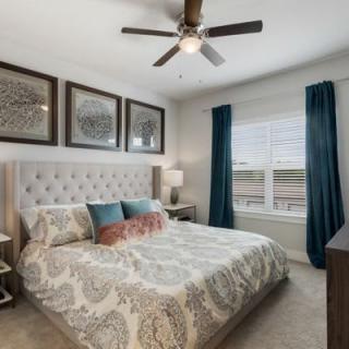 A cozy bedroom with a tufted headboard, patterned bedding, and blue curtains decorating the window.