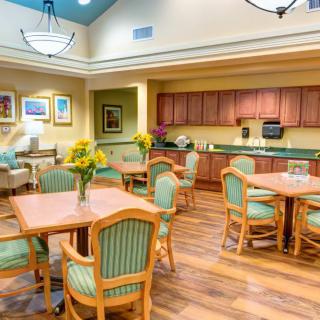 A brightly lit dining area with wooden tables and green cushioned chairs, featuring sunflowers as centerpieces and a kitchenette in the background.