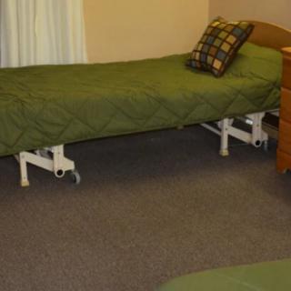 A simple bedroom with a single bed featuring green bedding and a plaid pillow, next to a wooden nightstand on a carpeted floor.