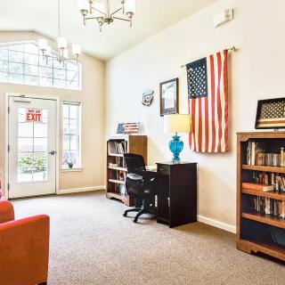 A brightly lit office interior featuring bookshelves, a desk with a chair and lamp, and an American flag on the wall.
