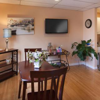 A cozy dining room with a wooden table and chairs, a wall-mounted television, and decorative elements such as a painting, lamp, and plant.