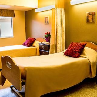 A brightly lit nursing home or hospital room with two beds covered in yellow bedspreads and red pillows.