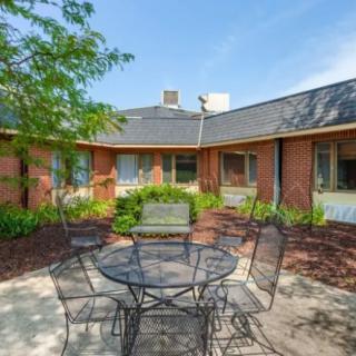 A tranquil courtyard with a metal outdoor table and chairs surrounded by greenery and a brick building.