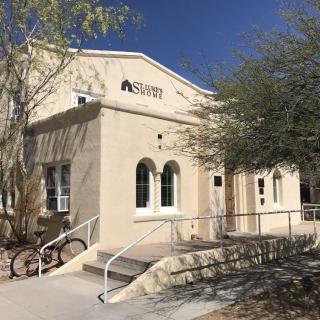 A small building in a desert setting with a bicycle parked outside and trees providing shade.