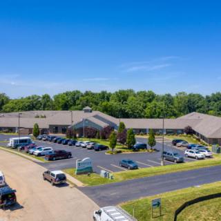 A large building with a parking lot filled with cars, surrounded by trees under a clear blue sky.