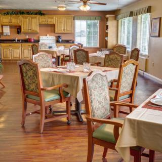A cozy dining room with neatly arranged tables and chairs, featuring wooden flooring and large windows with curtains.