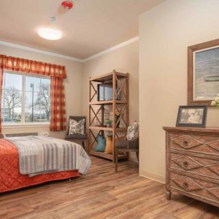 A cozy bedroom with wooden flooring, featuring a neatly made bed, a dresser with artwork on top, and a shelf by the window.