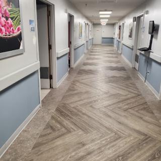 A well-lit interior hallway in a healthcare facility, featuring herringbone patterned flooring and decorative wall art.