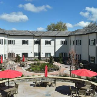 A view of a courtyard with tables and red umbrellas surrounded by a two-story building under a blue sky with clouds.