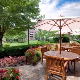 A beautiful outdoor patio area with wooden tables and chairs under umbrellas, surrounded by lush greenery and blooming flowers.