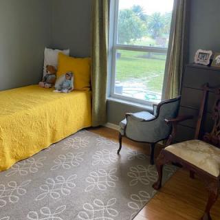A cozy bedroom featuring a single bed with yellow bedding, an armchair by the window, and a patterned rug.