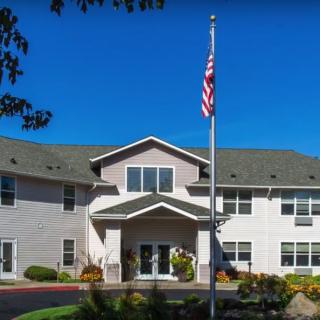 A two-story building with an American flag in front, surrounded by landscaped greenery under a clear blue sky.