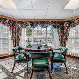 A traditional dining area with a round table and green upholstered chairs, surrounded by large windows and colorful curtains.