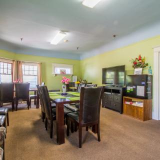 A well-lit dining room with a large table and several chairs, surrounded by windows and decorated shelves.