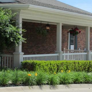 A brick house with a covered porch, surrounded by landscaped shrubs and hanging plants.