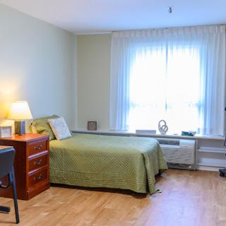 A cozy and minimalist bedroom featuring a green bedspread, a wooden dresser, and ample natural light.
