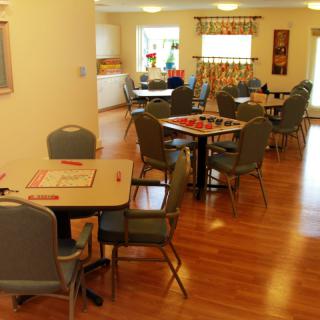 A cozy community dining area with multiple tables and chairs, featuring hardwood floors and large windows.