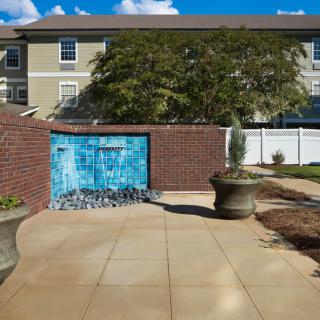 A tranquil outdoor area featuring a small fountain set against a brick wall, surrounded by planters and paved walkways.