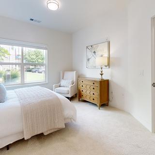 A bright and cozy bedroom with a bed, dresser, lamp, and chair near a large window.