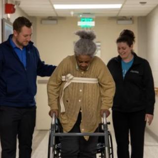 An elderly person using a walker is assisted by two healthcare professionals in a hallway.
