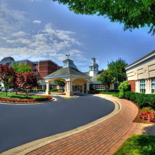 A scenic view of a building with a circular driveway, surrounded by trees and lush landscaping under a partly cloudy sky.