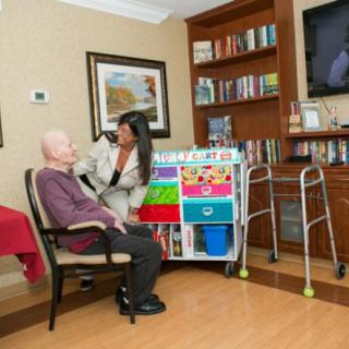 A healthcare worker interacting with an elderly person in a nursing home, surrounded by bookshelves and a walker.