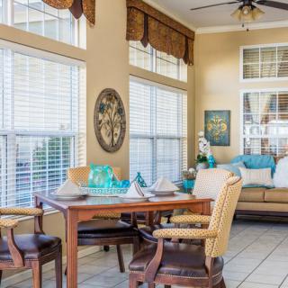A cozy dining and living area filled with natural light from large windows, featuring a wooden table and chairs surrounded by decorative elements.
