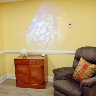 A cozy living room corner with a comfortable armchair beside a wooden cabinet, featuring a colorful projection on the yellow wall.
